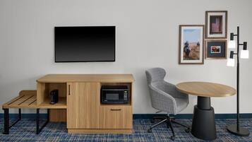 A modern hotel room with a flat-screen TV, a wooden cabinet with a microwave, a gray office chair, a round table, and framed pictures on the wall.
