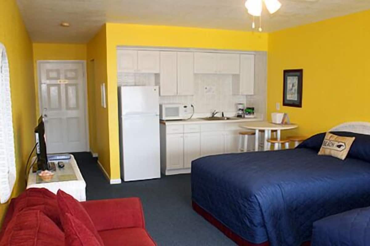A compact hotel room with a kitchenette, a single bed, a red chair, and a small table.