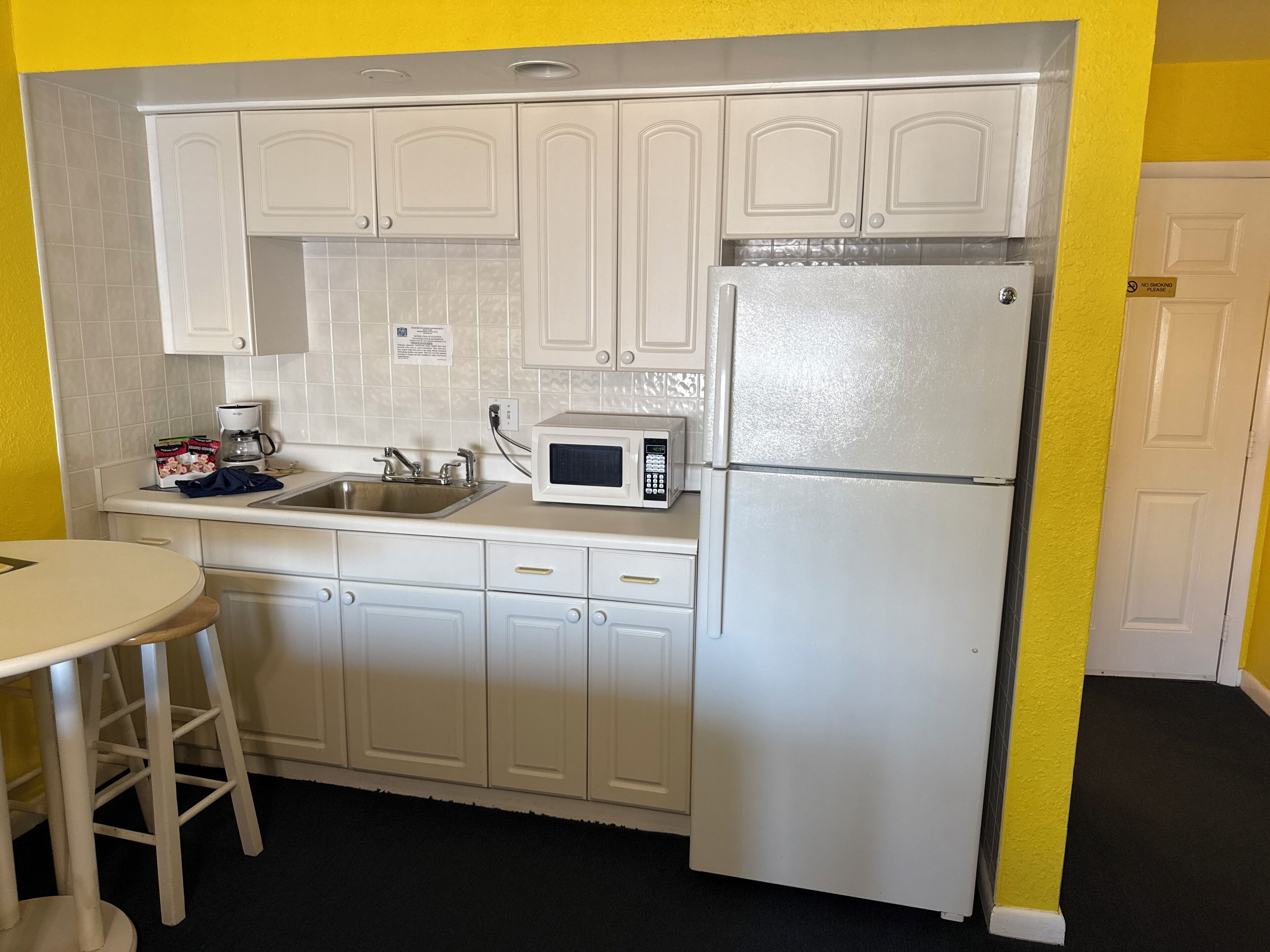 A compact kitchen with white cabinets, a microwave, a refrigerator, a sink, and a small round table with a stool.