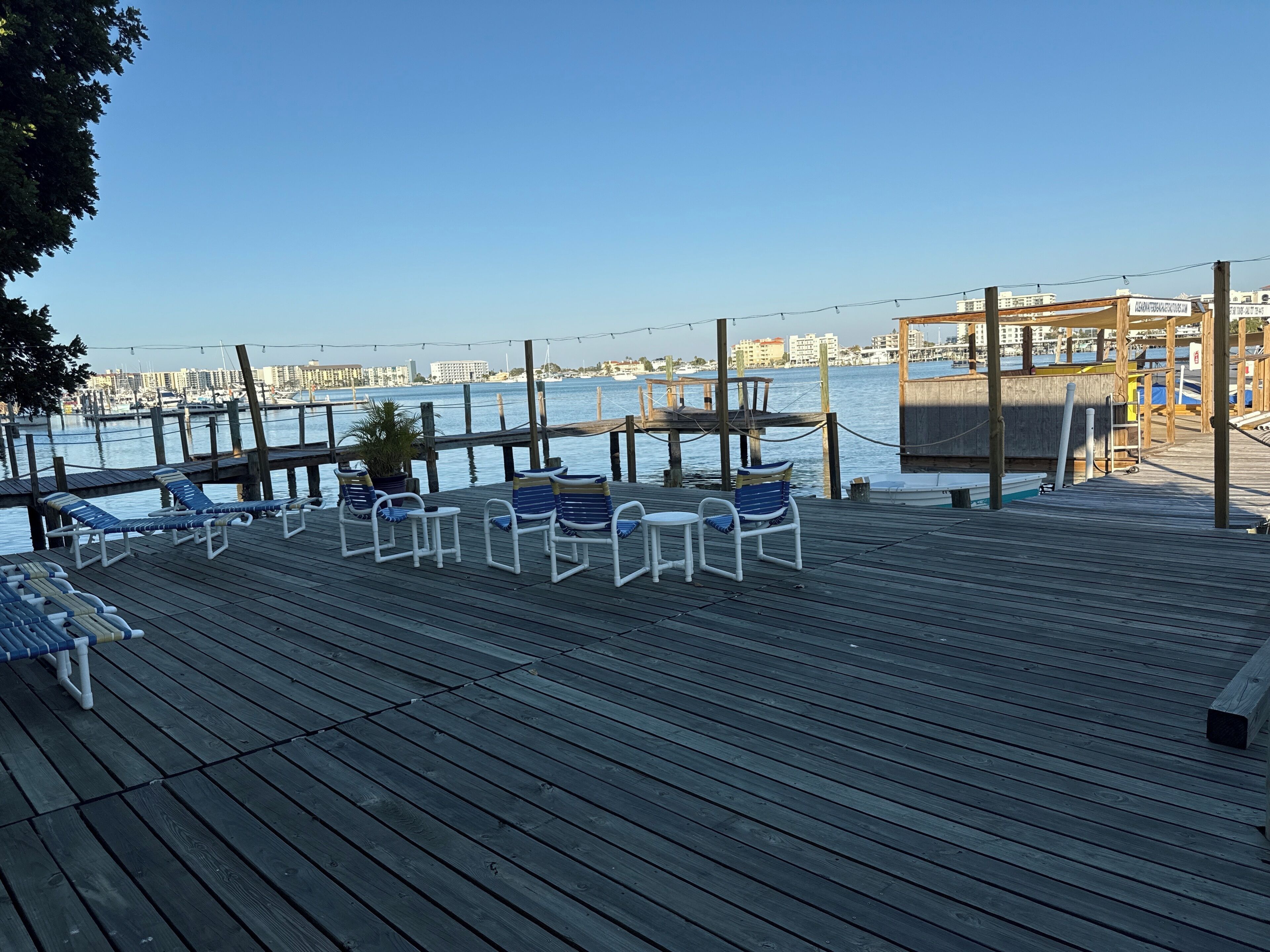 A wooden deck with lounge chairs and tables overlooking a marina with boats and buildings in the background.