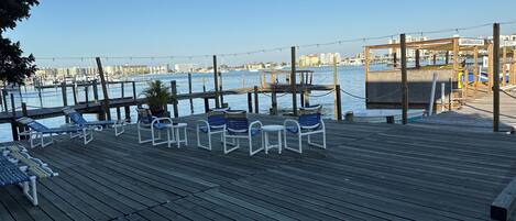 A wooden deck with lounge chairs and tables overlooking a marina with boats and buildings in the background.