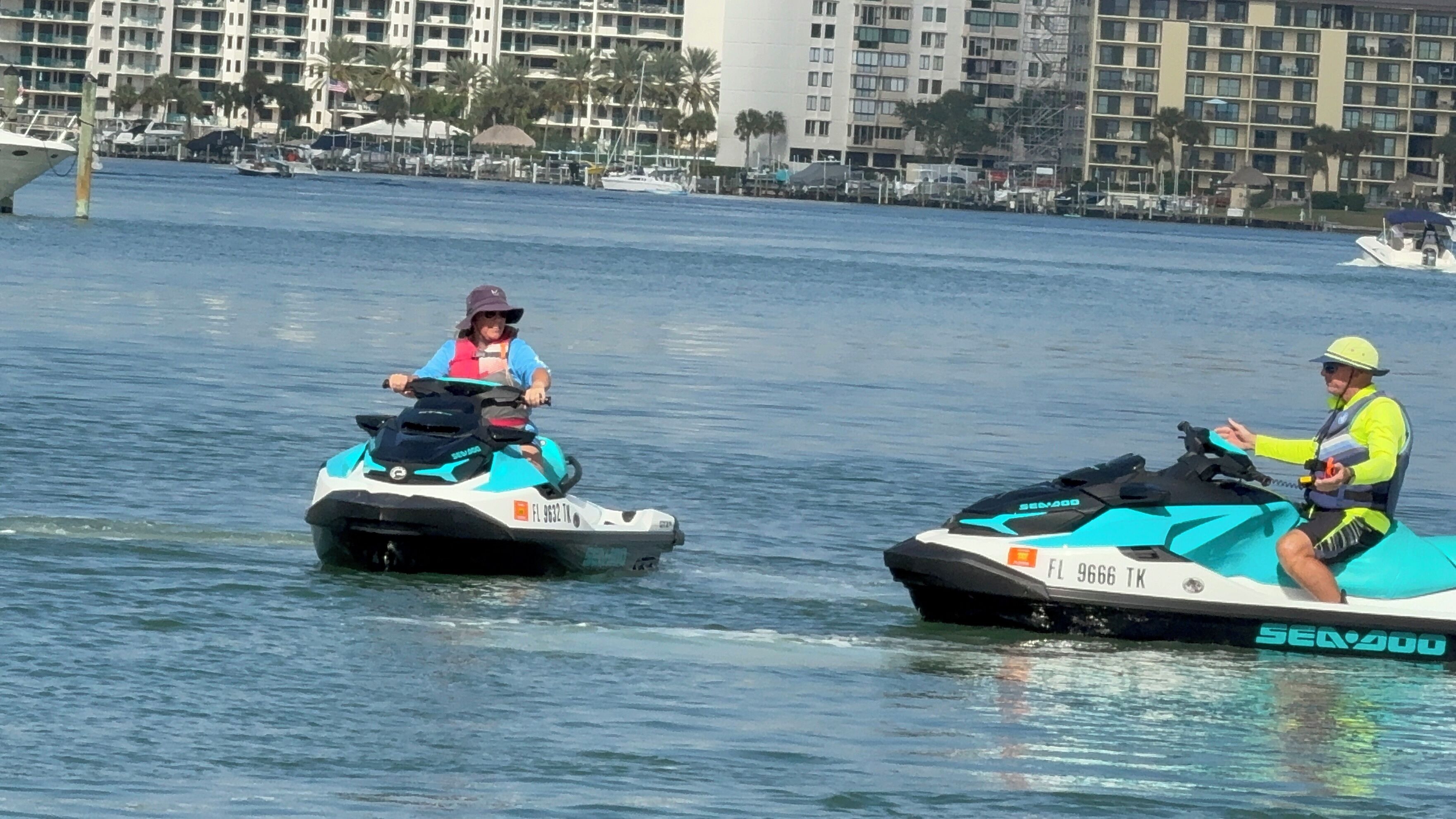 Twee personen op jetski's op het water, met een jachthaven en gebouwen op de achtergrond.