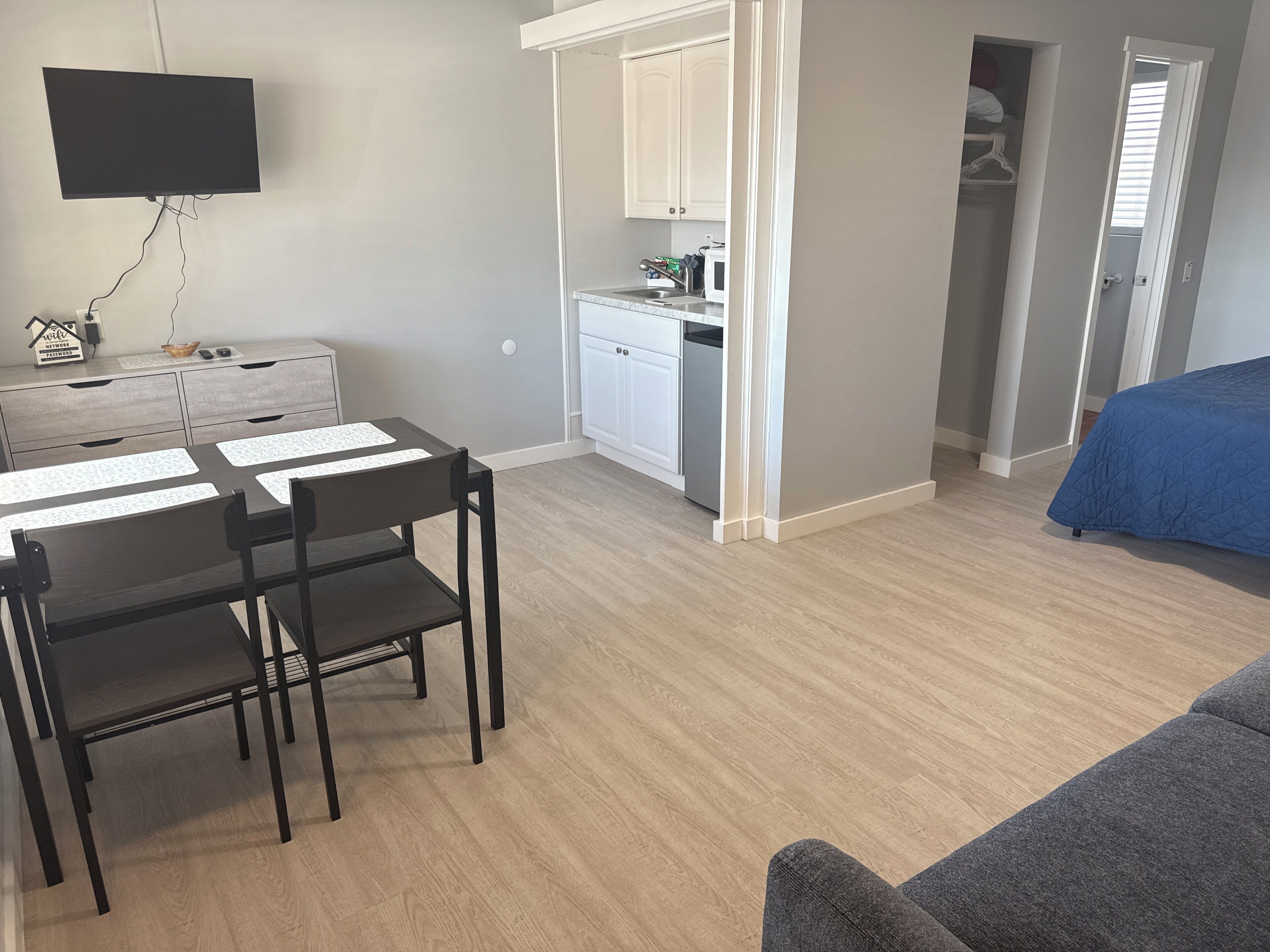 A compact room with a dining table, chairs, a bed, a TV, and a kitchenette.