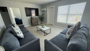 Ein Wohnzimmer mit zwei grauen Sofas, einem Couchtisch, einem Flachbildfernseher, einer Topfpflanze und einem Fenster mit Jalousien.