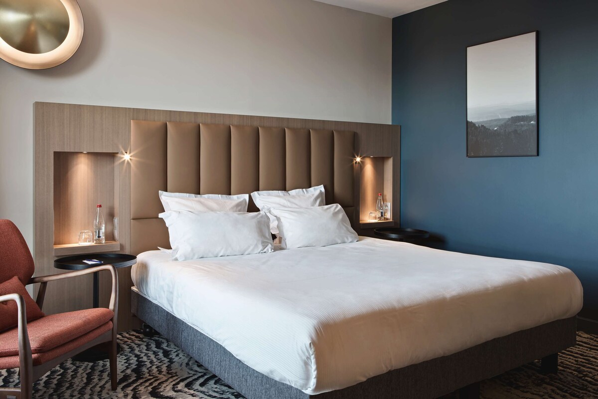 Une chambre d’hôtel moderne avec un grand lit, une tête de lit dotée d’un éclairage intégré, une table de chevet avec une bouteille et des verres, un fauteuil rouge et un tableau encadré au mur.