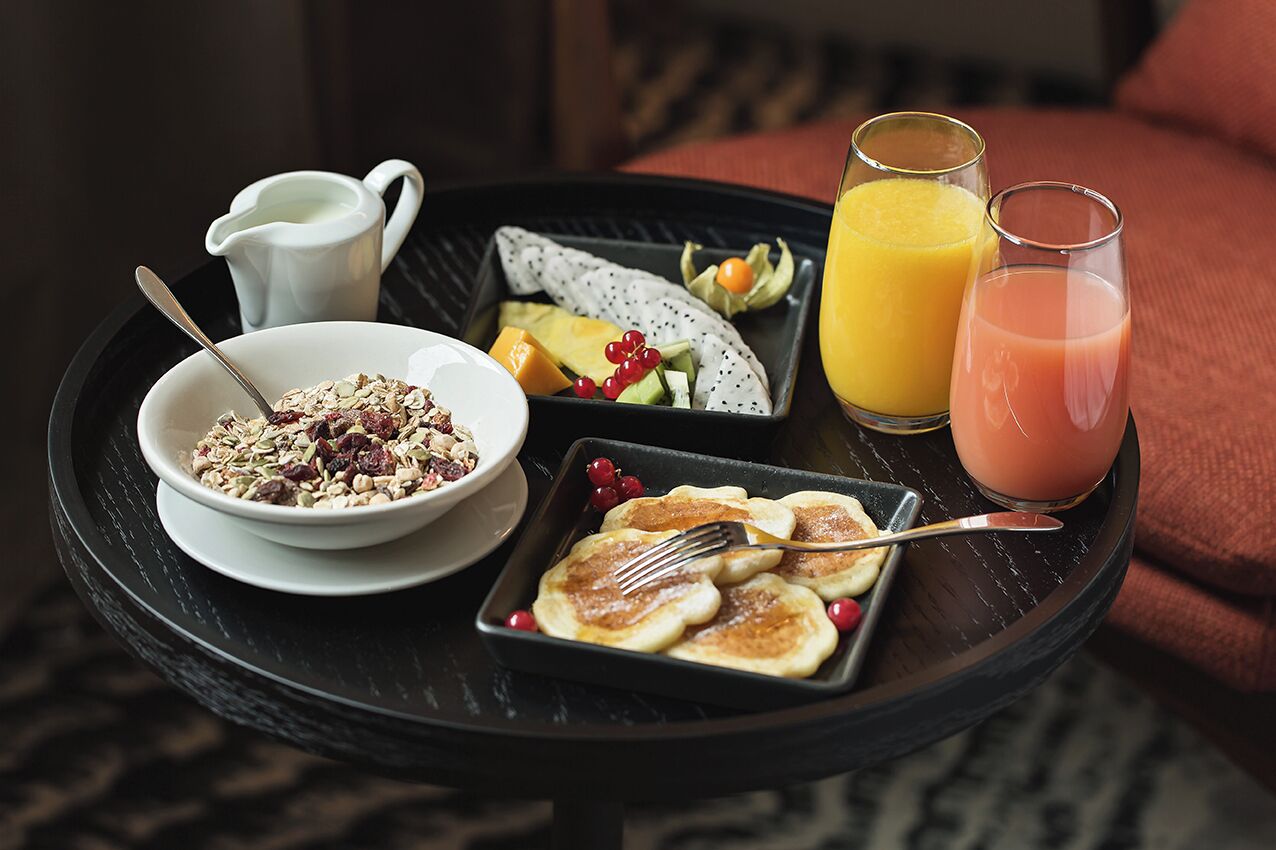 Una bandeja de desayuno con cereal, tortitas, fruta y dos vasos de zumo de naranja.