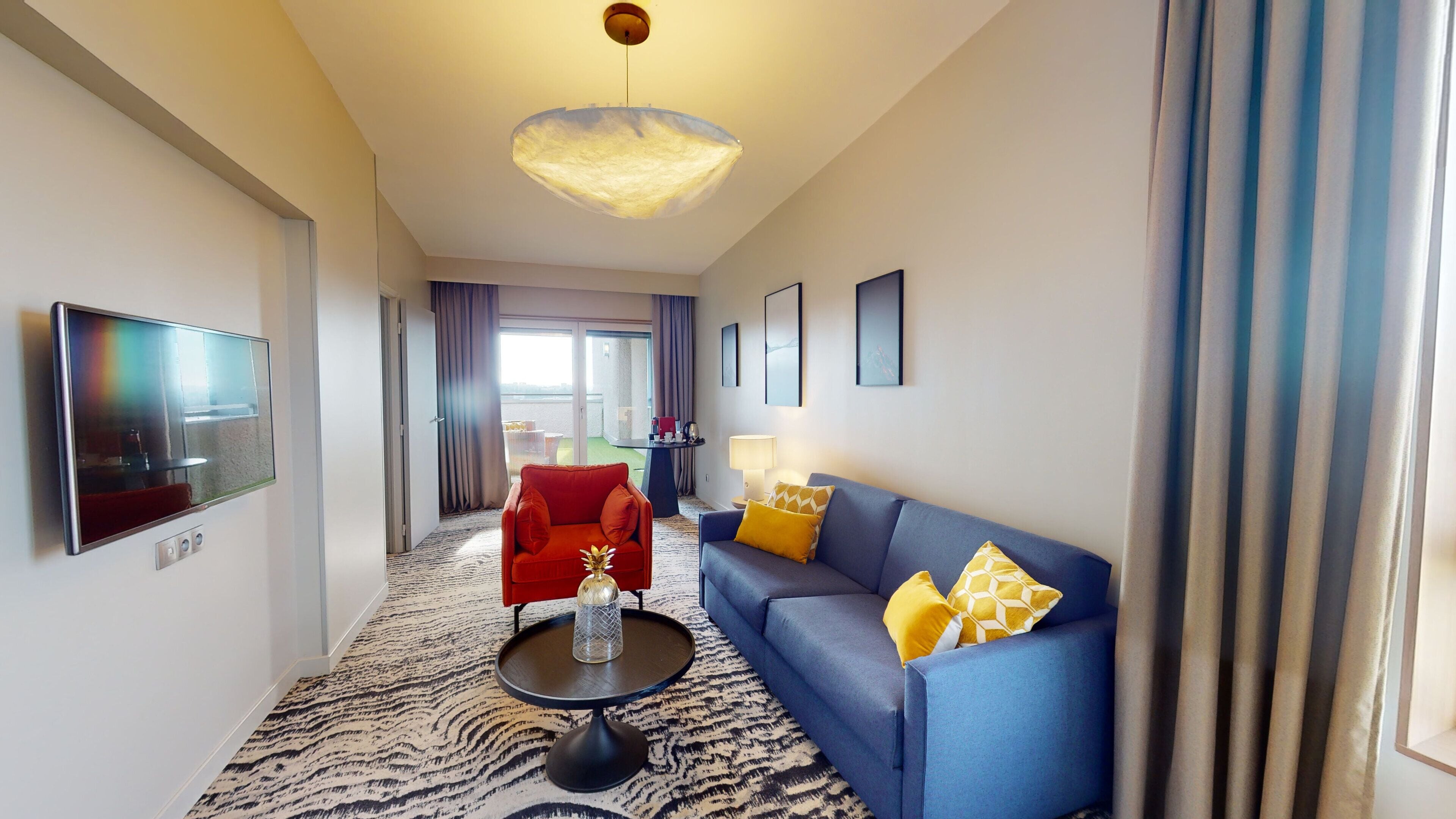 A modern hotel room with a flat-screen TV, a red armchair, a blue sofa, and a patterned carpet.