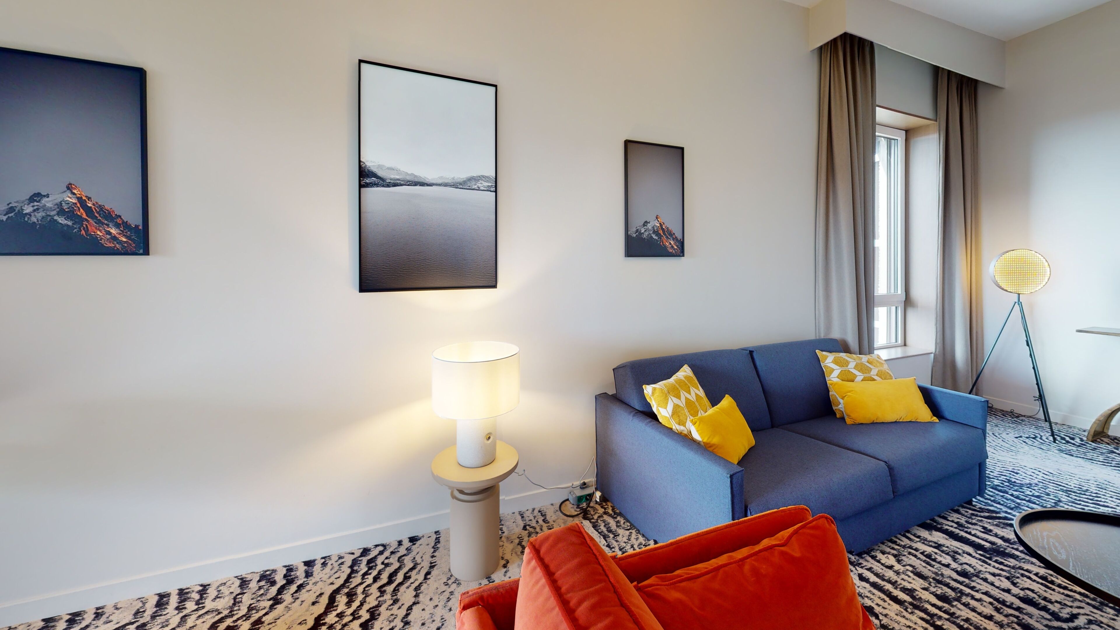 A modern hotel room with a blue sofa, yellow and orange cushions, a standing lamp, and framed artwork on the walls.