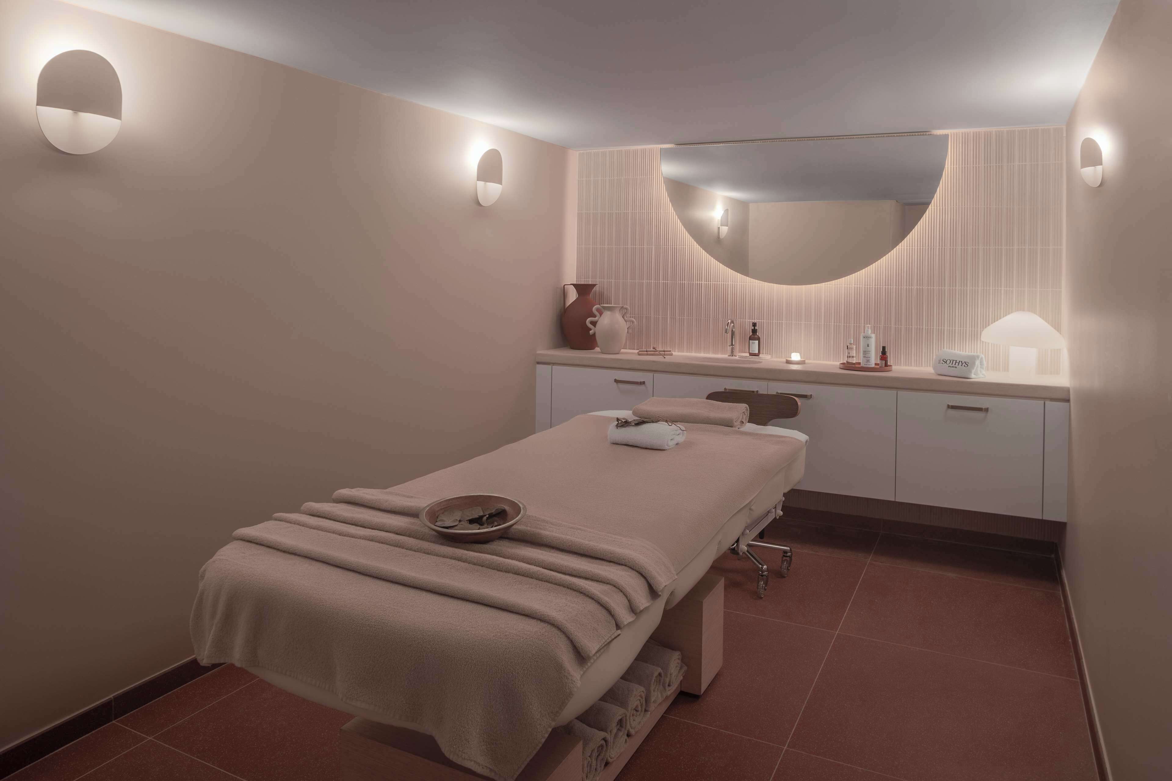 A modern spa room with a massage table, a large mirror, and a vanity with toiletries.