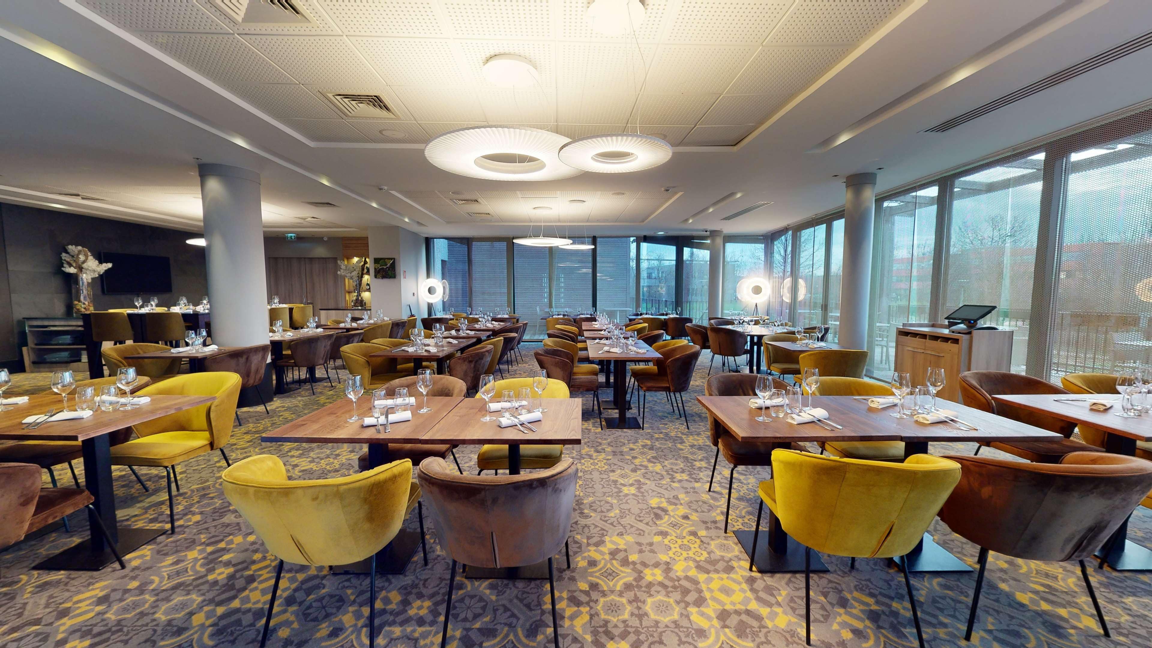 A spacious restaurant with multiple dining tables, yellow and brown chairs, and large windows providing natural light.