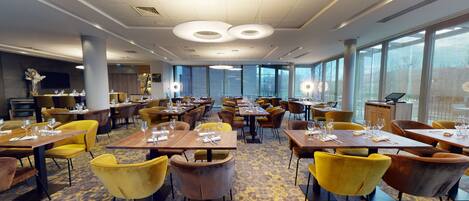 A spacious restaurant with multiple dining tables, yellow and brown chairs, and large windows providing natural light.