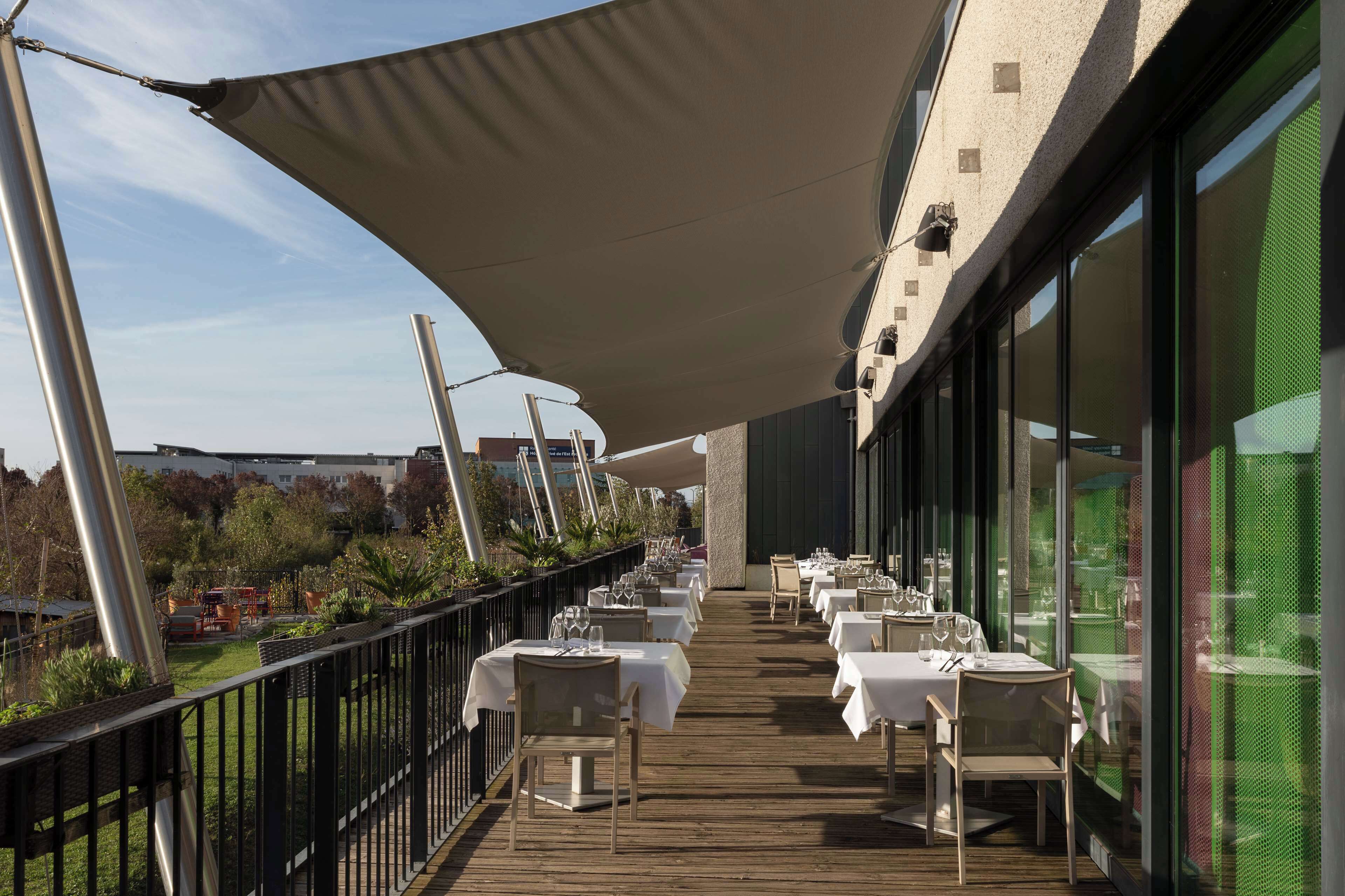 An outdoor dining area with tables and chairs, a wooden deck, and a large canopy providing shade.