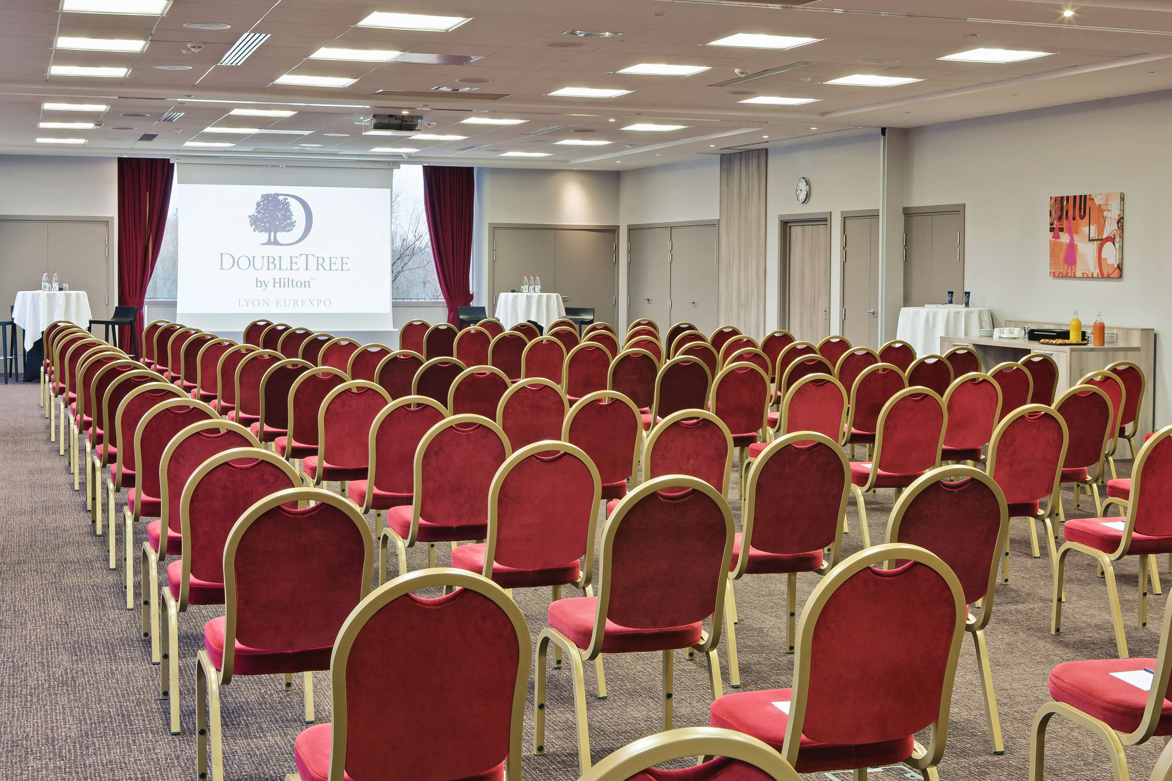 A conference room with rows of red chairs, a projection screen displaying "DoubleTree by Hilton Lyon Expo", and a clock on the wall.