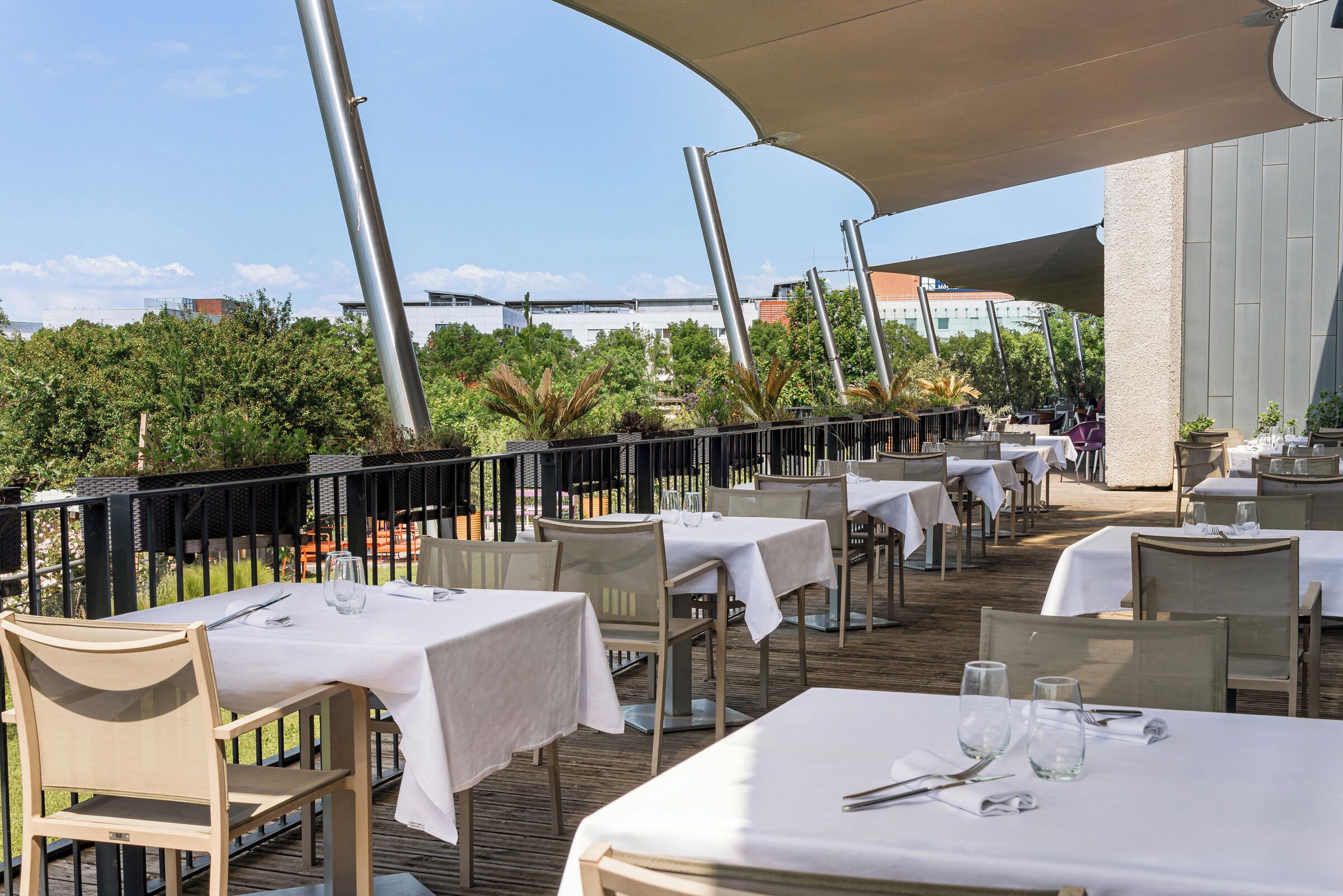 An outdoor dining area with tables and chairs, surrounded by greenery and buildings in the background.