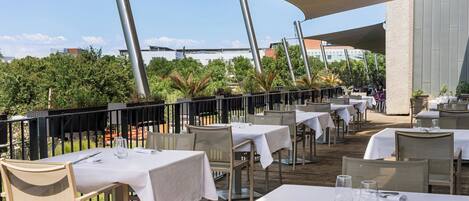 An outdoor dining area with tables and chairs, surrounded by greenery and buildings in the background.