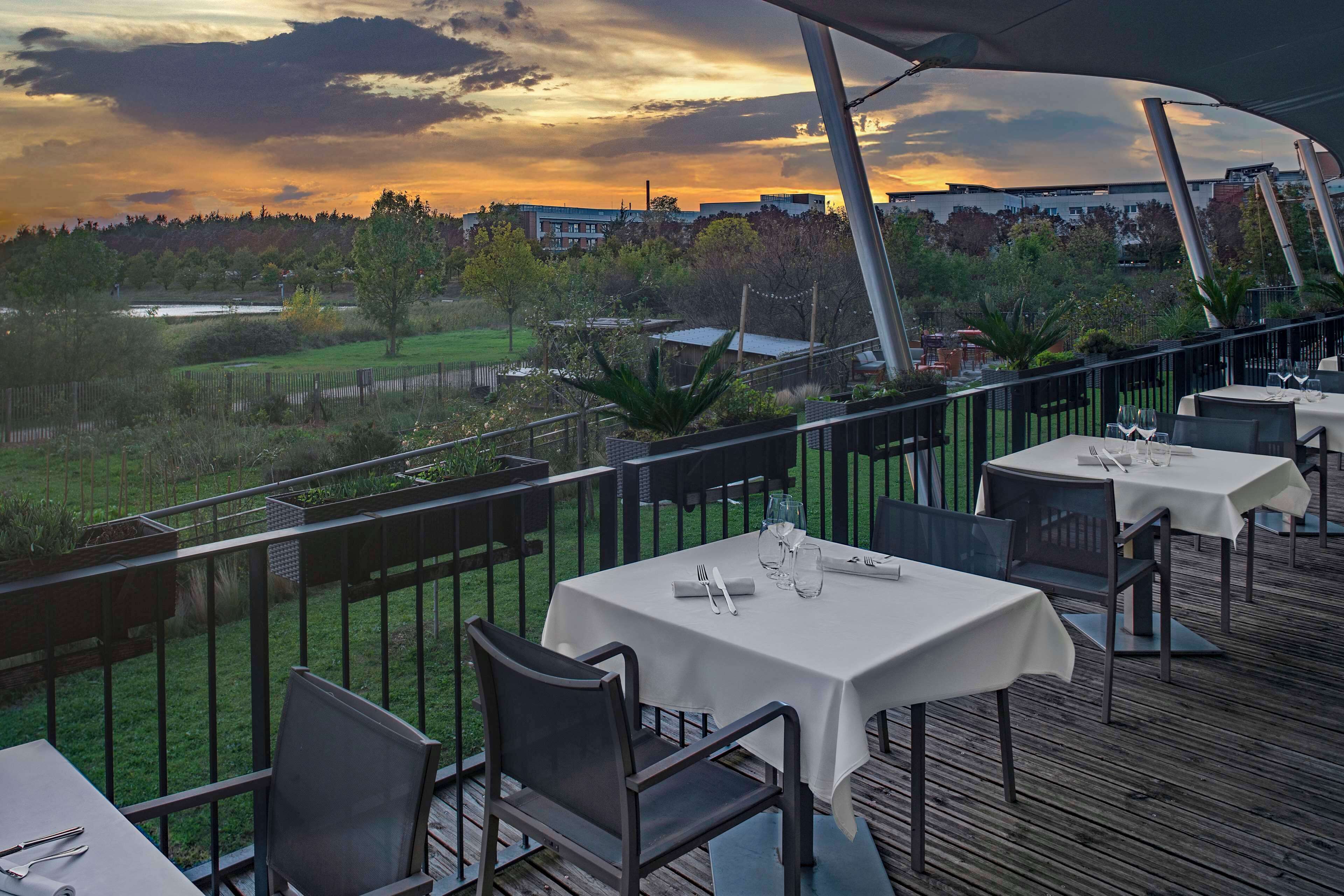 An outdoor dining area with tables and chairs, overlooking a grassy area and a body of water during sunset.