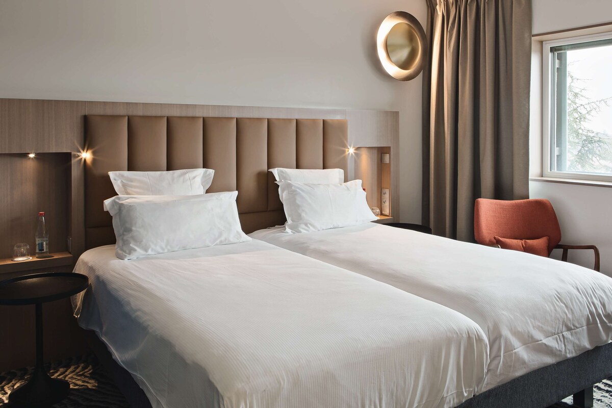 Une chambre d’hôtel avec deux lits, un miroir rond, une fenêtre avec des rideaux et une petite table sur laquelle se trouve une bouteille.