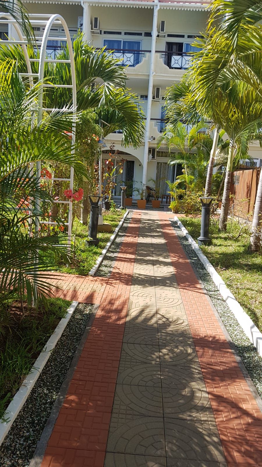 A pathway leading to a building with a white facade and blue accents, surrounded by palm trees and greenery.