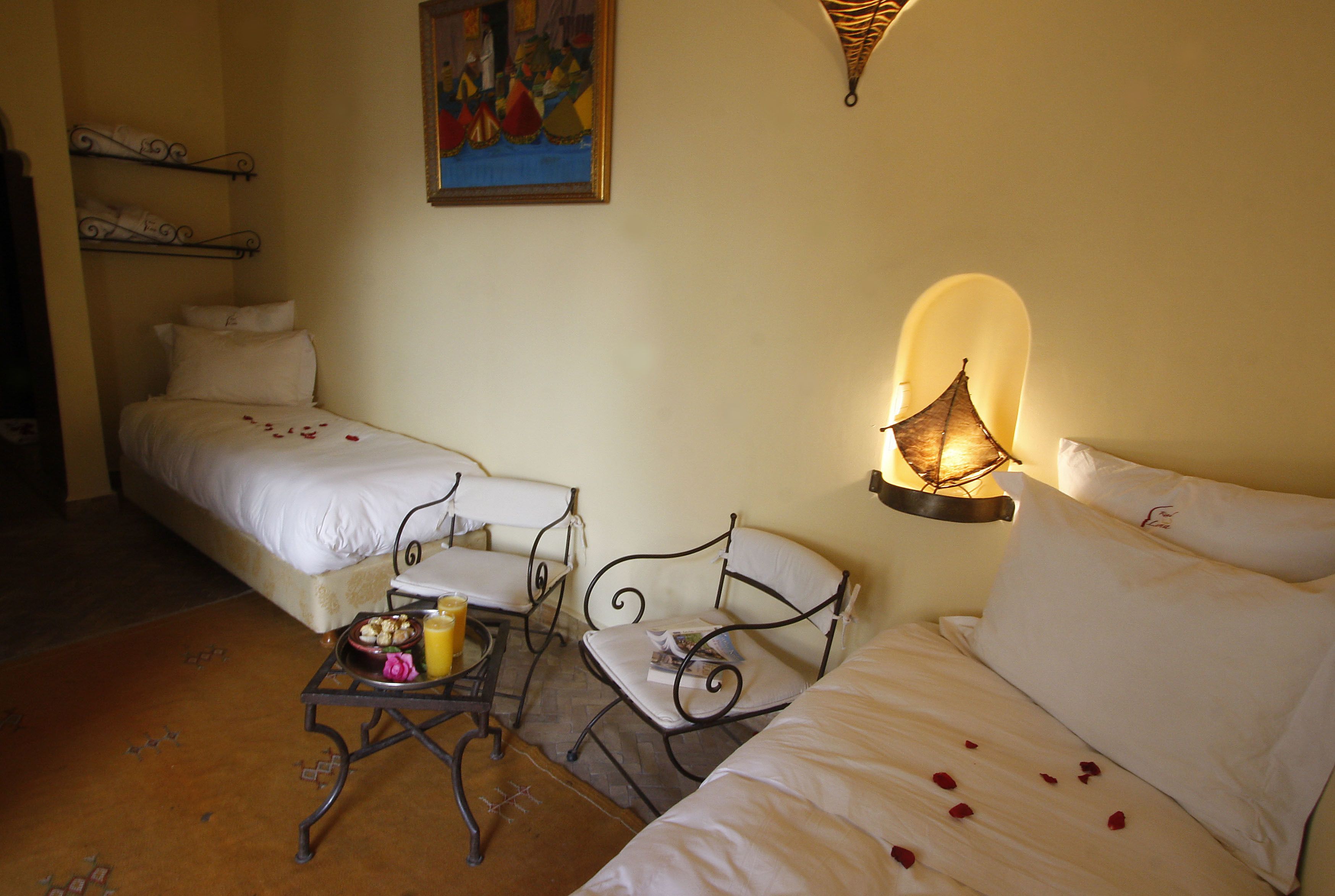 A bedroom with two beds, a small table with a book and a glass of juice, a chair, and a wall-mounted lamp.