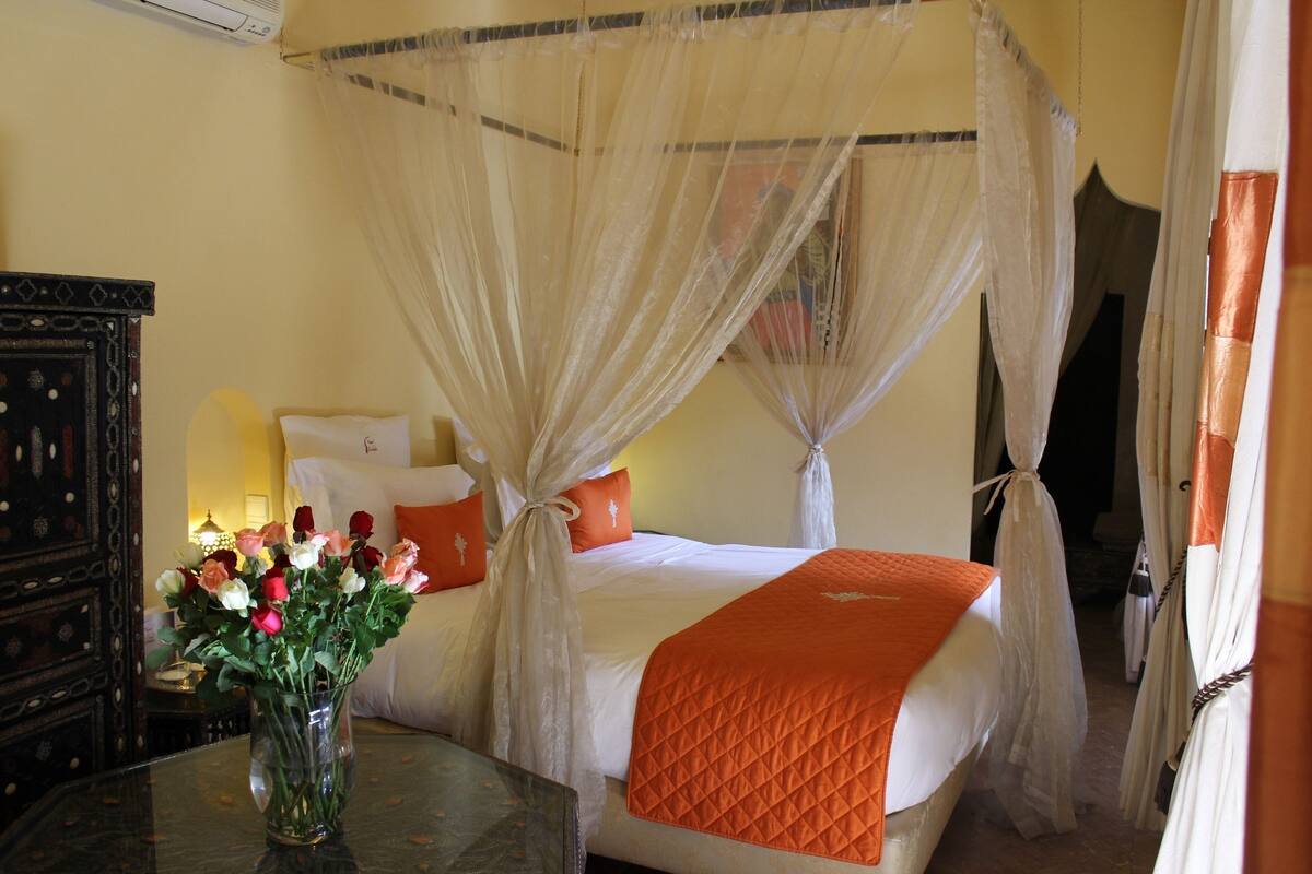 Un lit à baldaquin avec une couvre-lit blanche et un plaid matelassé orange, un vase de fleurs sur une table et un meuble en bois foncé.