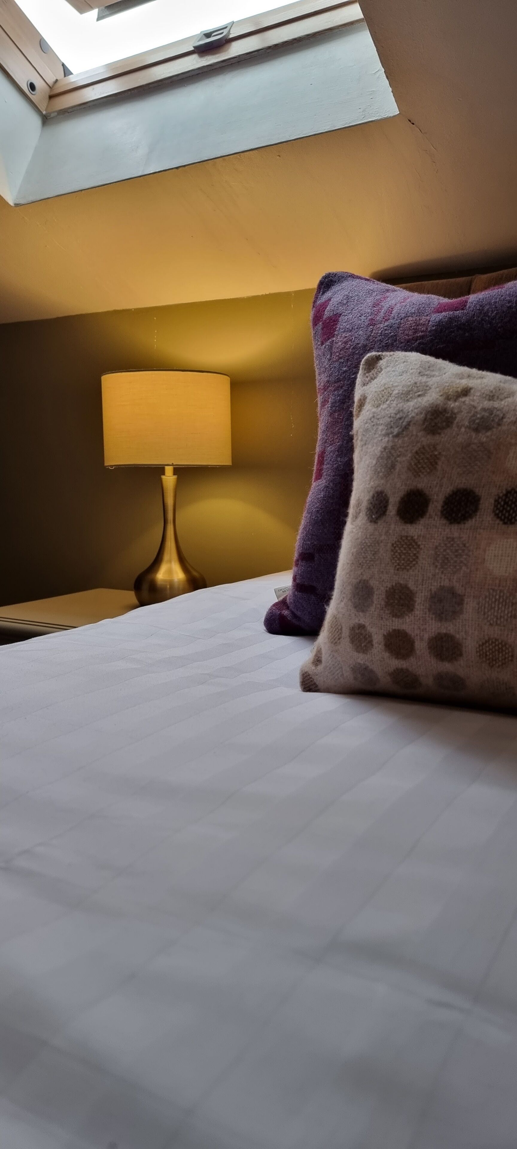 Un lit bien fait, avec une couette blanche, une lampe sur une table de chevet et deux coussins à motifs.