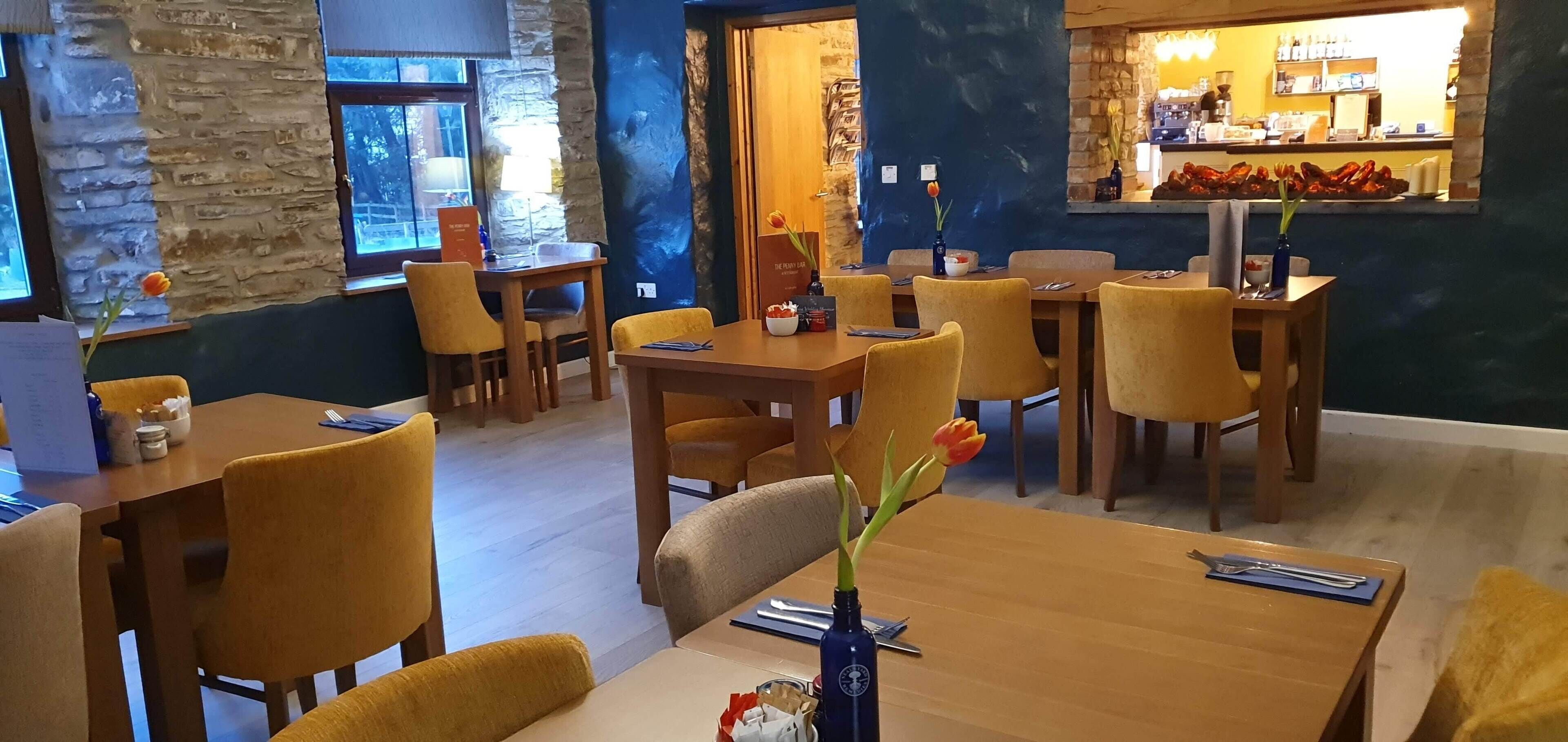 A restaurant with wooden tables and yellow chairs, stone walls, and a bar area in the background.