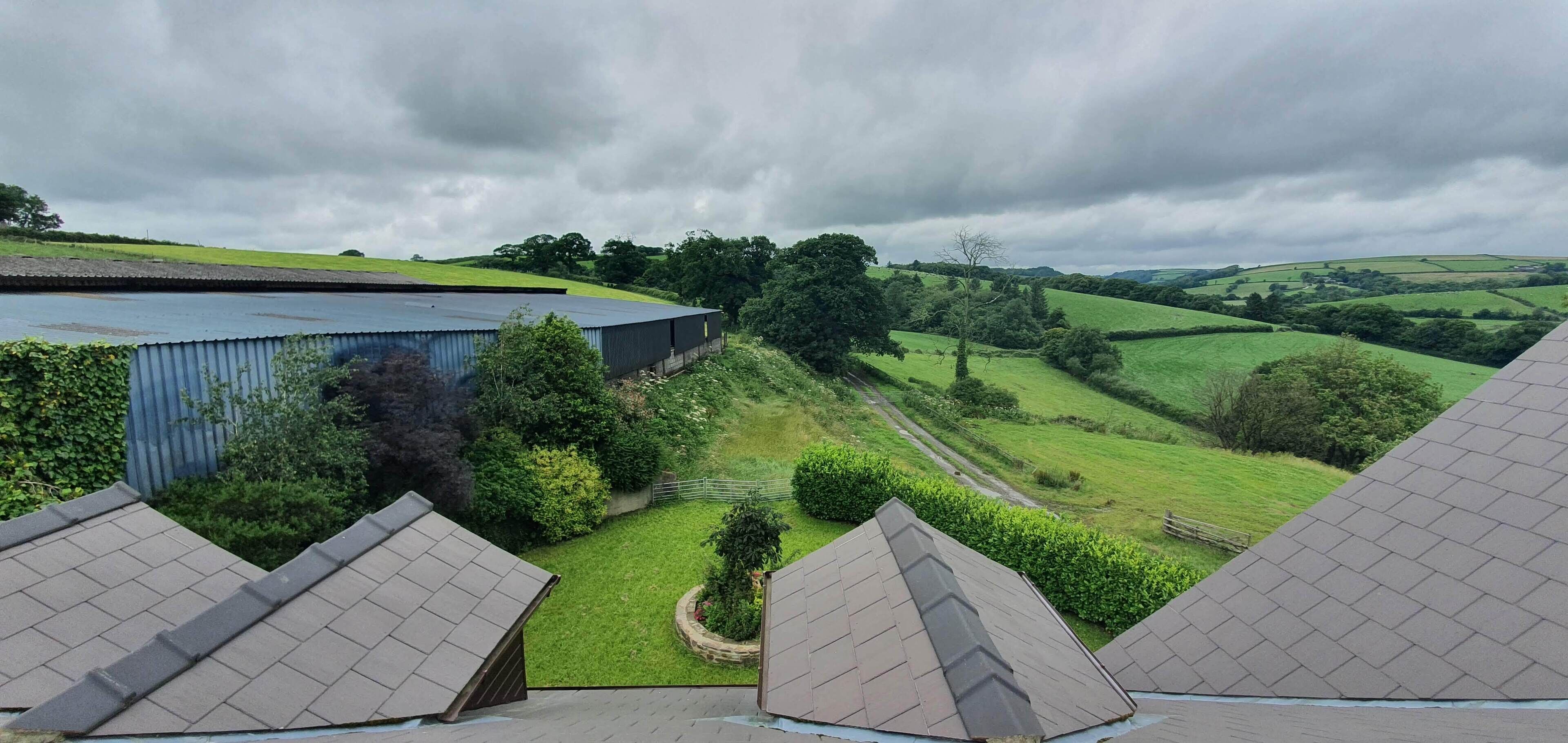 A view of a building with a sloped roof, surrounded by green fields and trees under a cloudy sky.