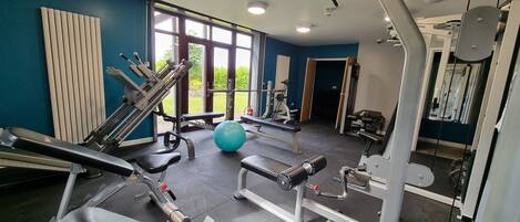 Un gimnasio con diversos equipos de ejercicio, amplias ventanas y vistas al verdor exterior.