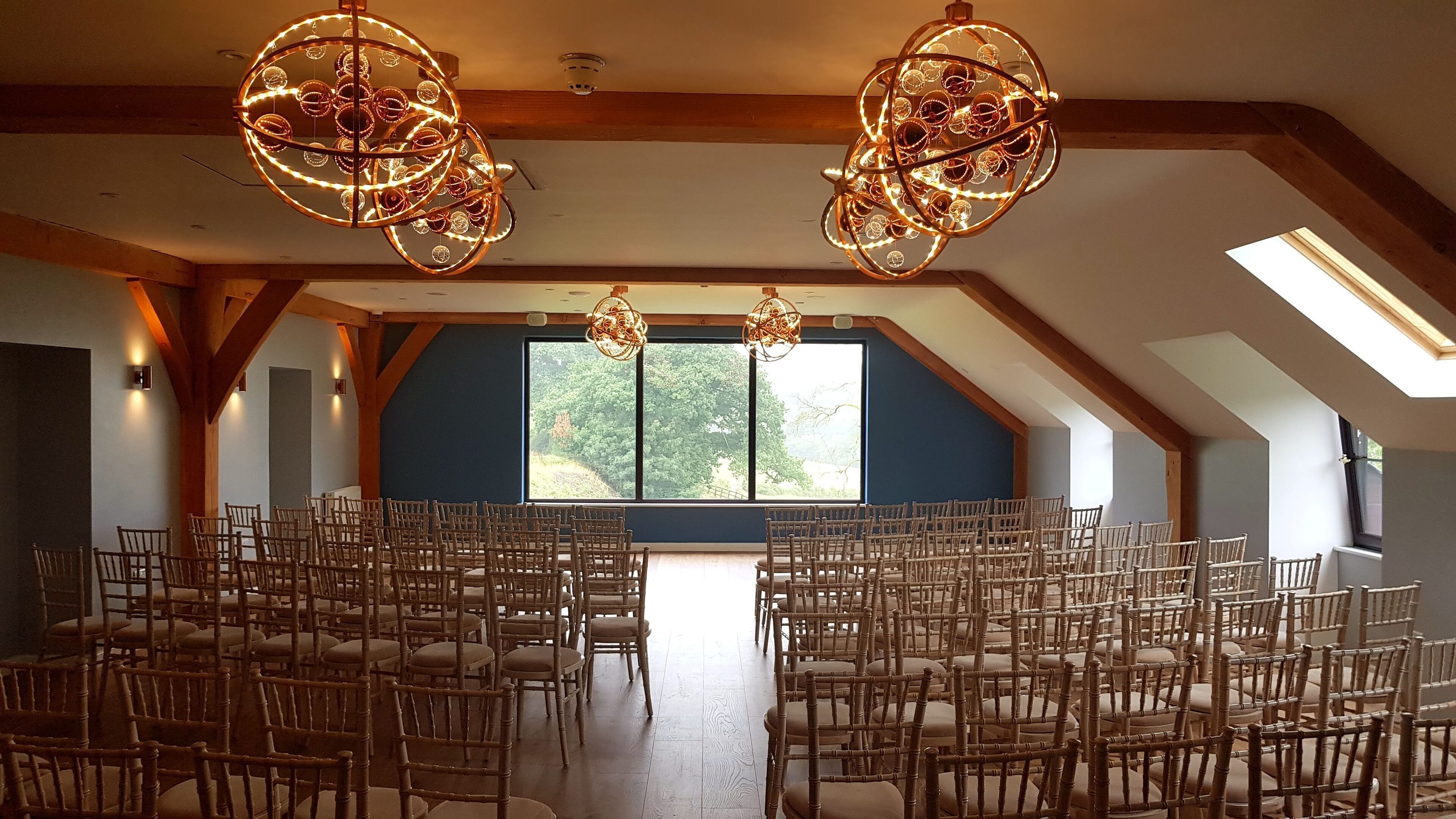 Ruime kamer met een hoge plafond, houten balken en een groot raam met uitzicht op bomen. Rijen stoelen staan klaar als zitplaatsen.
