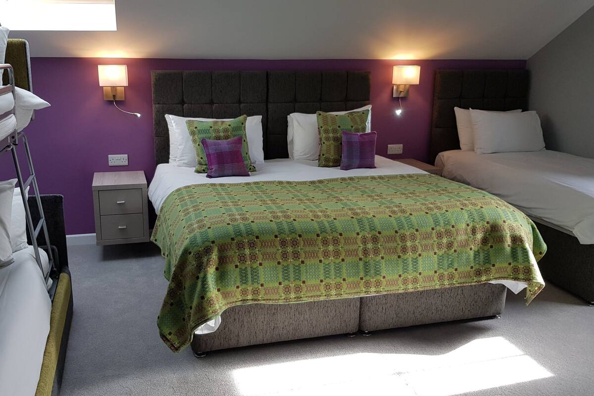 A room with a bunk bed, a single bed with a patterned bedspread, two bedside tables with lamps, and a purple accent wall.