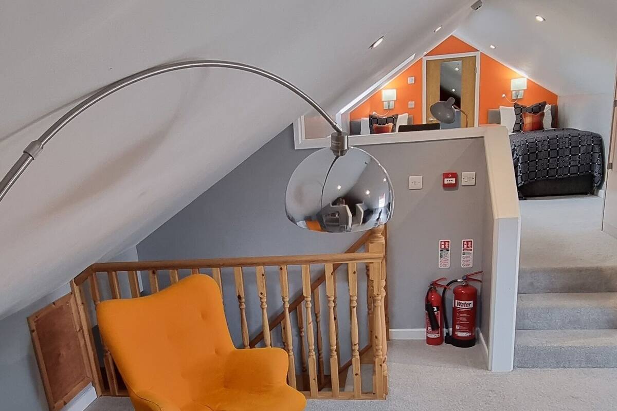 A modern loft bedroom with a sloped ceiling, a round table, orange chairs, a bed with a black and white patterned bedspread, and a wooden chair.