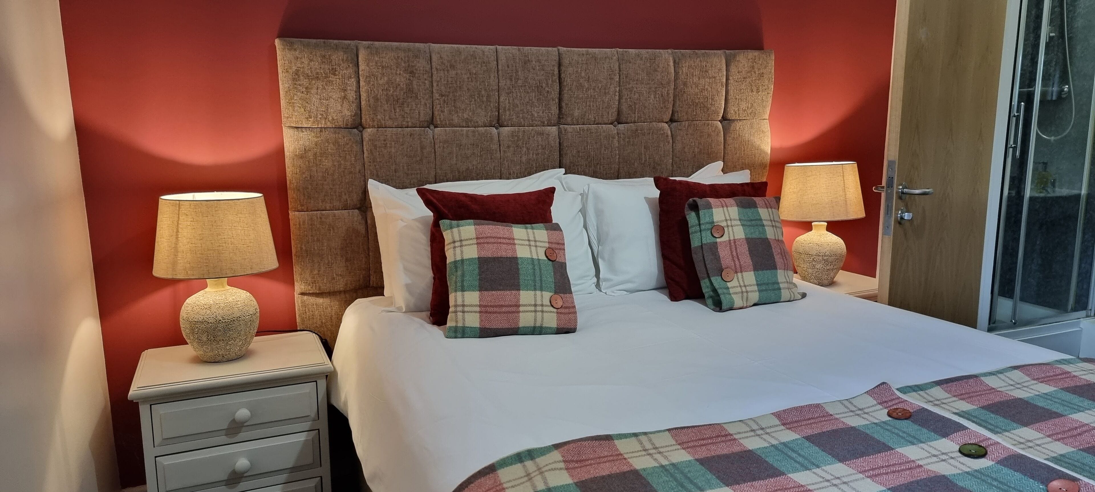 A bedroom with a tufted headboard, a bed with plaid pillows, two bedside lamps, and a glass shower door.