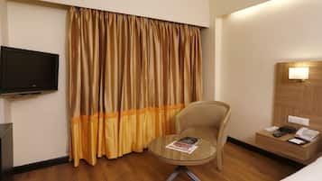 A hotel room with a television, a round glass table with a magazine, a chair, a bedside table with a phone, and a headboard with a light.