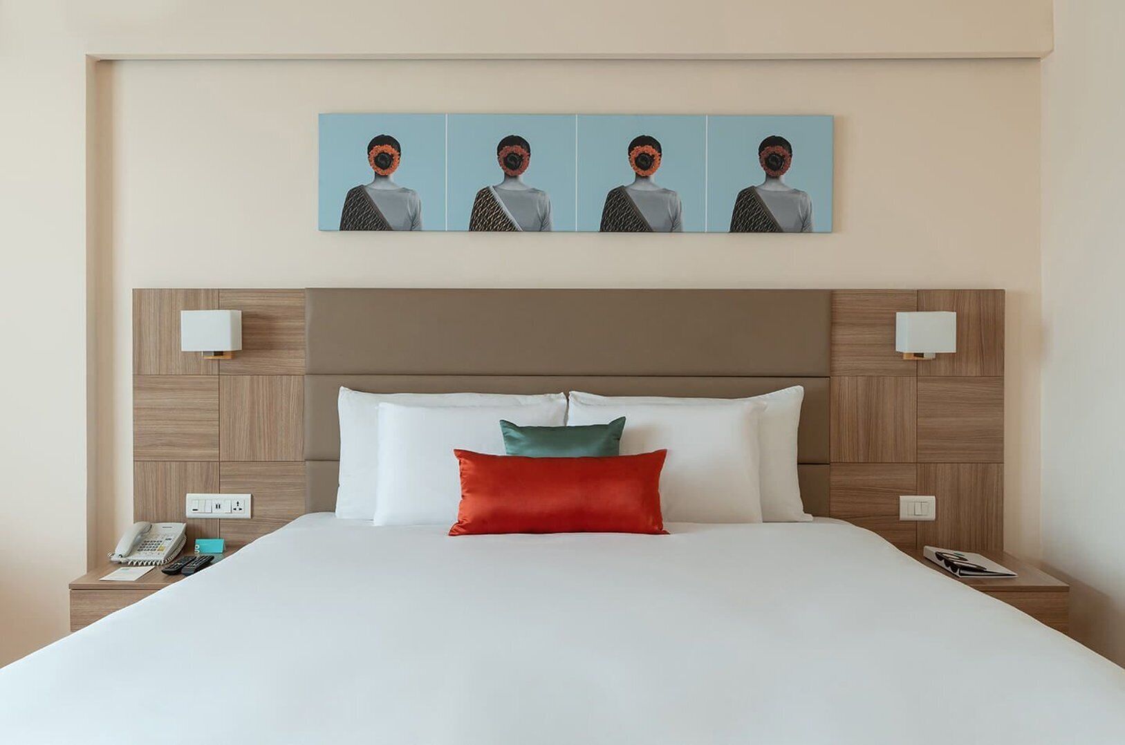 A hotel room with a large bed, a headboard with a light fixture, a wall-mounted artwork, and a bedside table with a phone and a remote control.