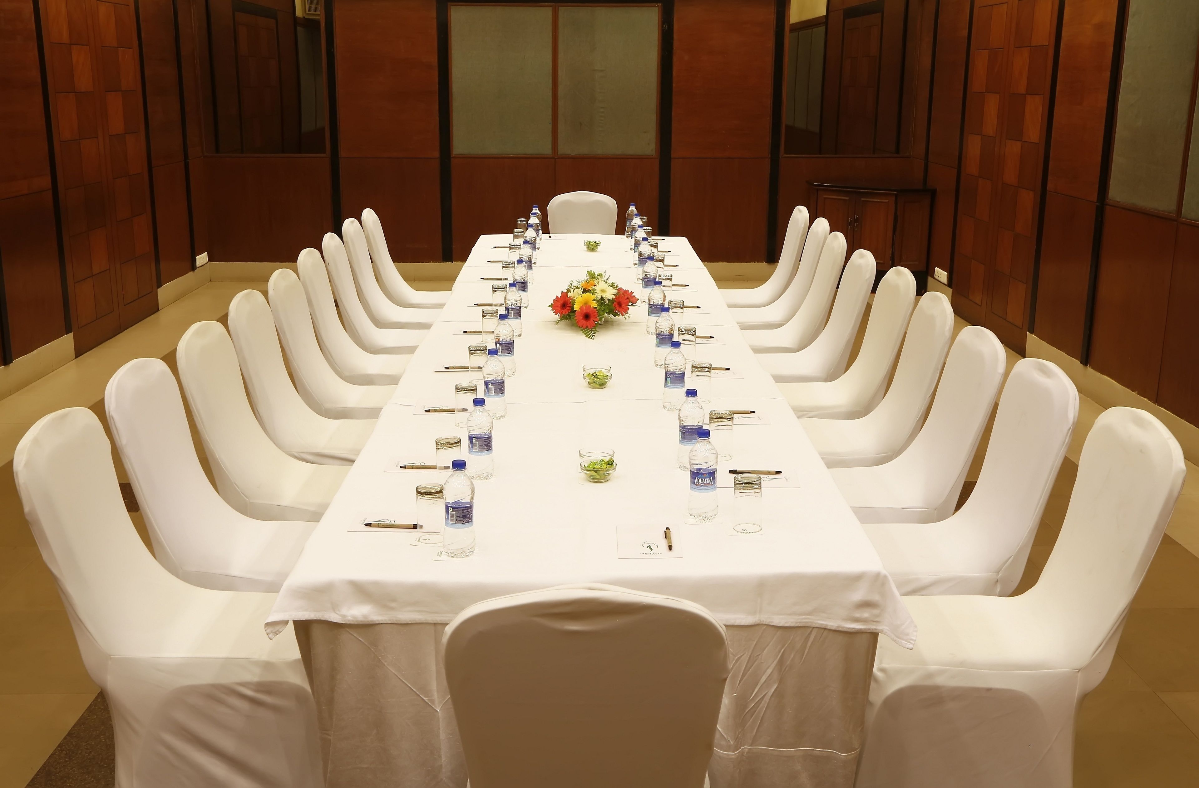 Un salón de conferencias con una larga mesa dispuesta para una reunión, rodeado de sillas blancas, con botellas de agua y un centro de mesa floral.
