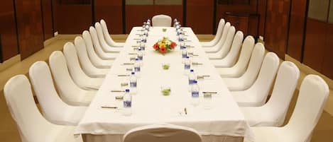 Un salón de conferencias con una larga mesa dispuesta para una reunión, rodeado de sillas blancas, con botellas de agua y un centro de mesa floral.