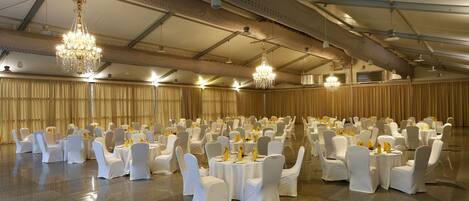 A banquet hall with round tables and white chairs, chandeliers, and a high ceiling.