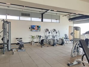 Un gimnasio con varios equipos de ejercicio, incluyendo cintas de correr y máquinas de pesas, y un televisor montado en la pared.