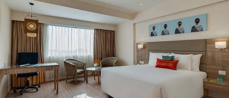Habitación de hotel con cama, escritorio con computadora, silla, una mesita y ventana con cortinas.