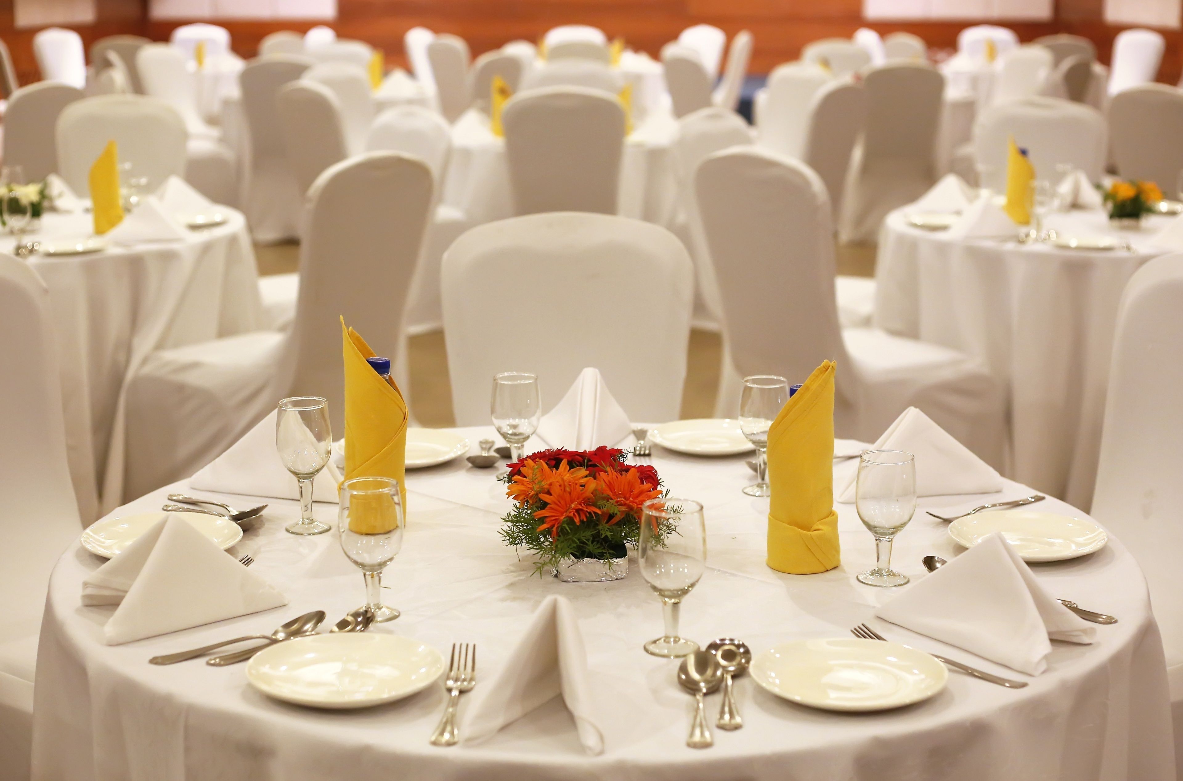 A banquet hall with round tables set for dining, each with white tablecloths, silverware, and yellow napkins.