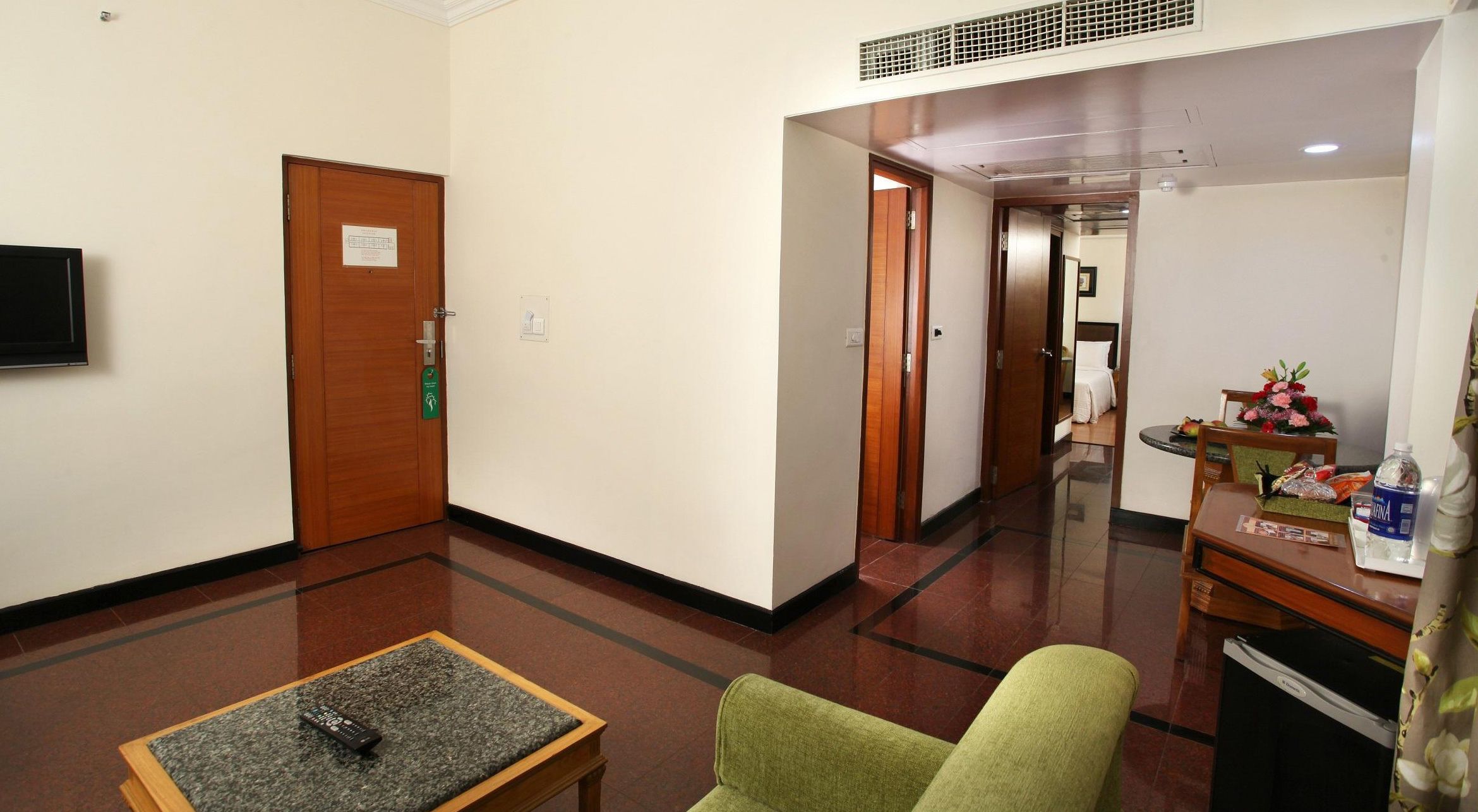 Un pasillo de hotel con una puerta de madera, una silla verde, una mesita con un control remoto y un minibar con una botella de agua.