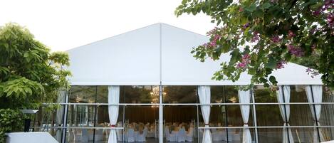 A modern building with a glass facade, white curtains, and a seating area with tables and chairs.