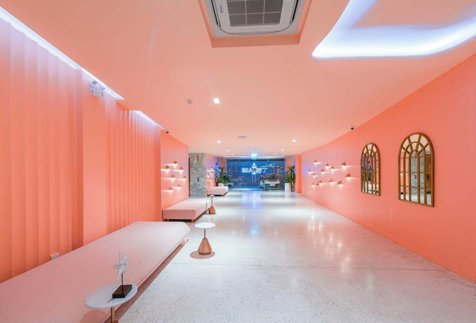 Un moderno vestíbulo de hotel con una decoración en tonos rosados, que incluye una recepción, zonas de estar y apliques decorativos en la pared.