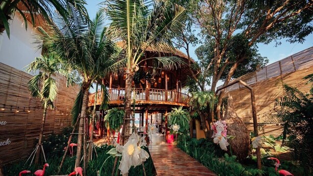 Un complexe hôtelier tropical doté d’une structure en bois, d’un toit de chaume et d’un sentier bordé de plantes tropicales et de flamants roses.