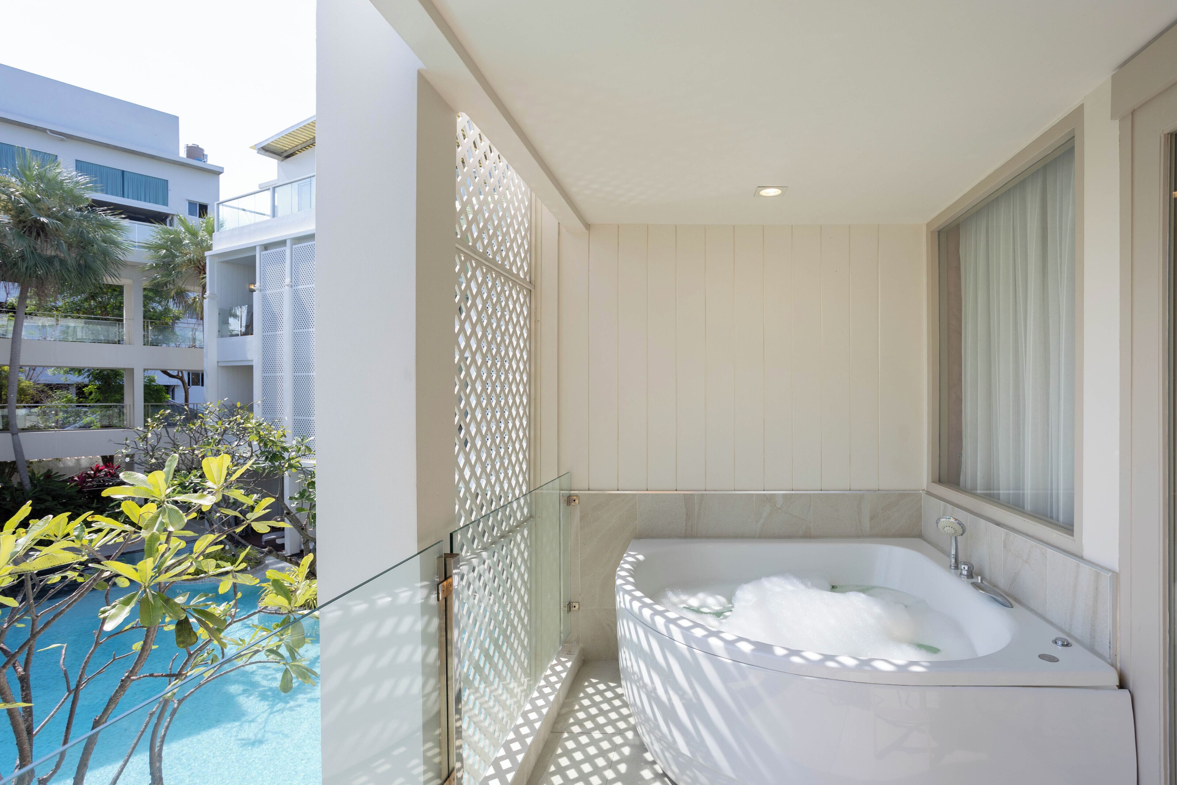 Un baño moderno con una bañera independiente, separador de vidrio y vistas a una piscina y follaje.