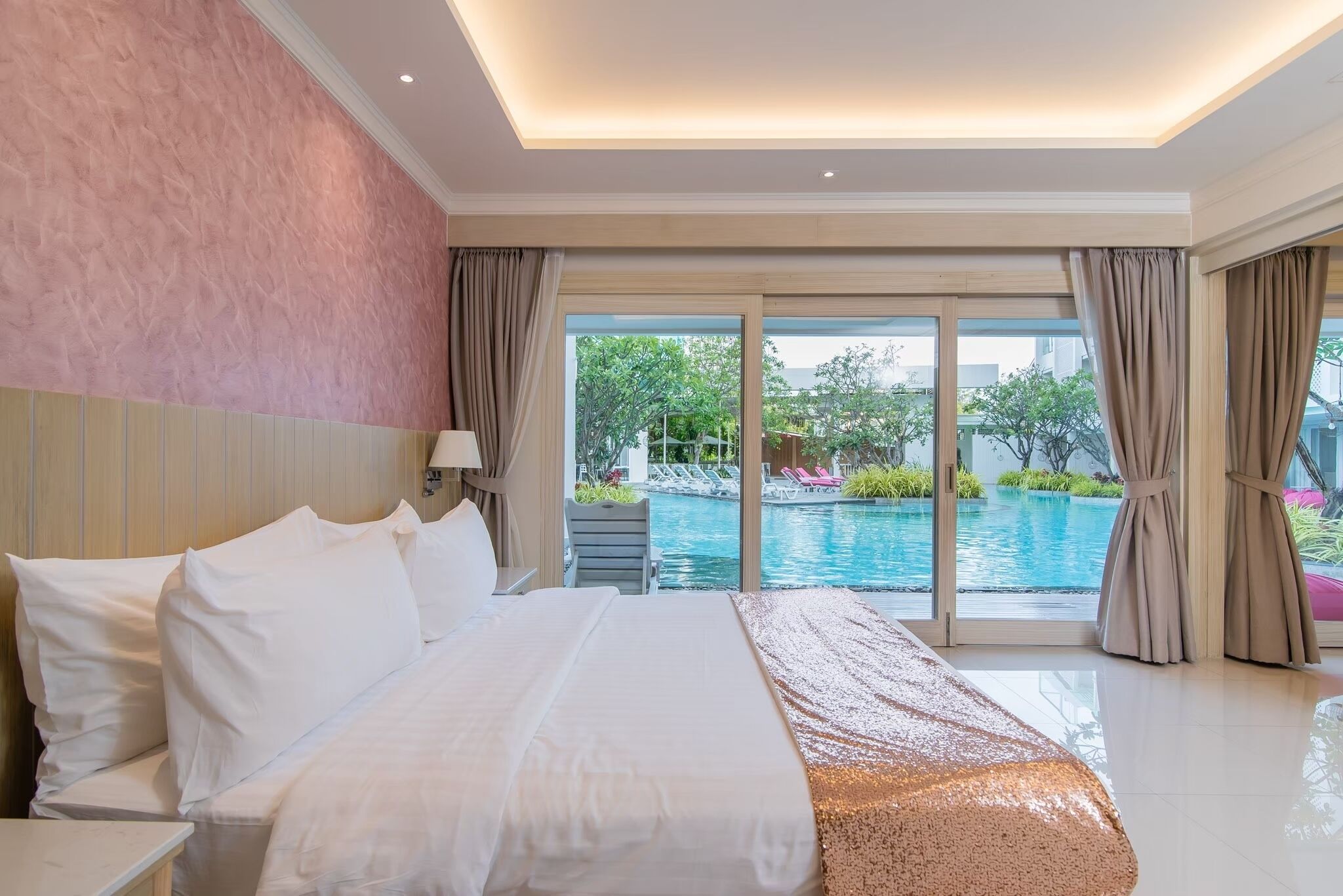 Une chambre d’hôtel avec un lit, une table de chevet, une lampe, une vue sur une piscine et un balcon avec des rideaux.