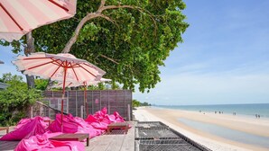 Une terrasse au bord de la plage, avec des chaises longues en bois, des coussins roses et des parasols à rayures.