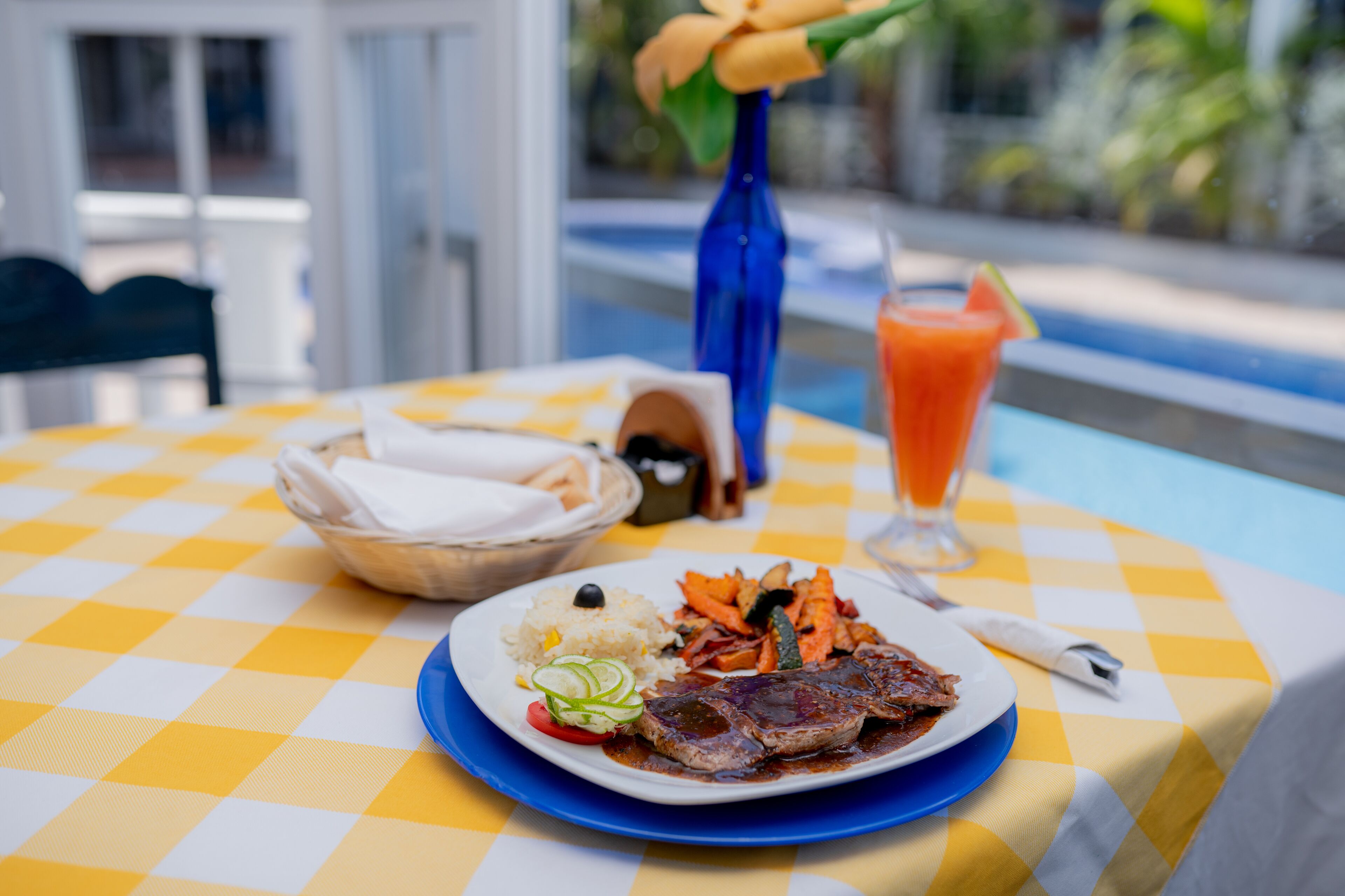 A dining table with a checkered tablecloth, a plate of food, a drink, and a vase with flowers.