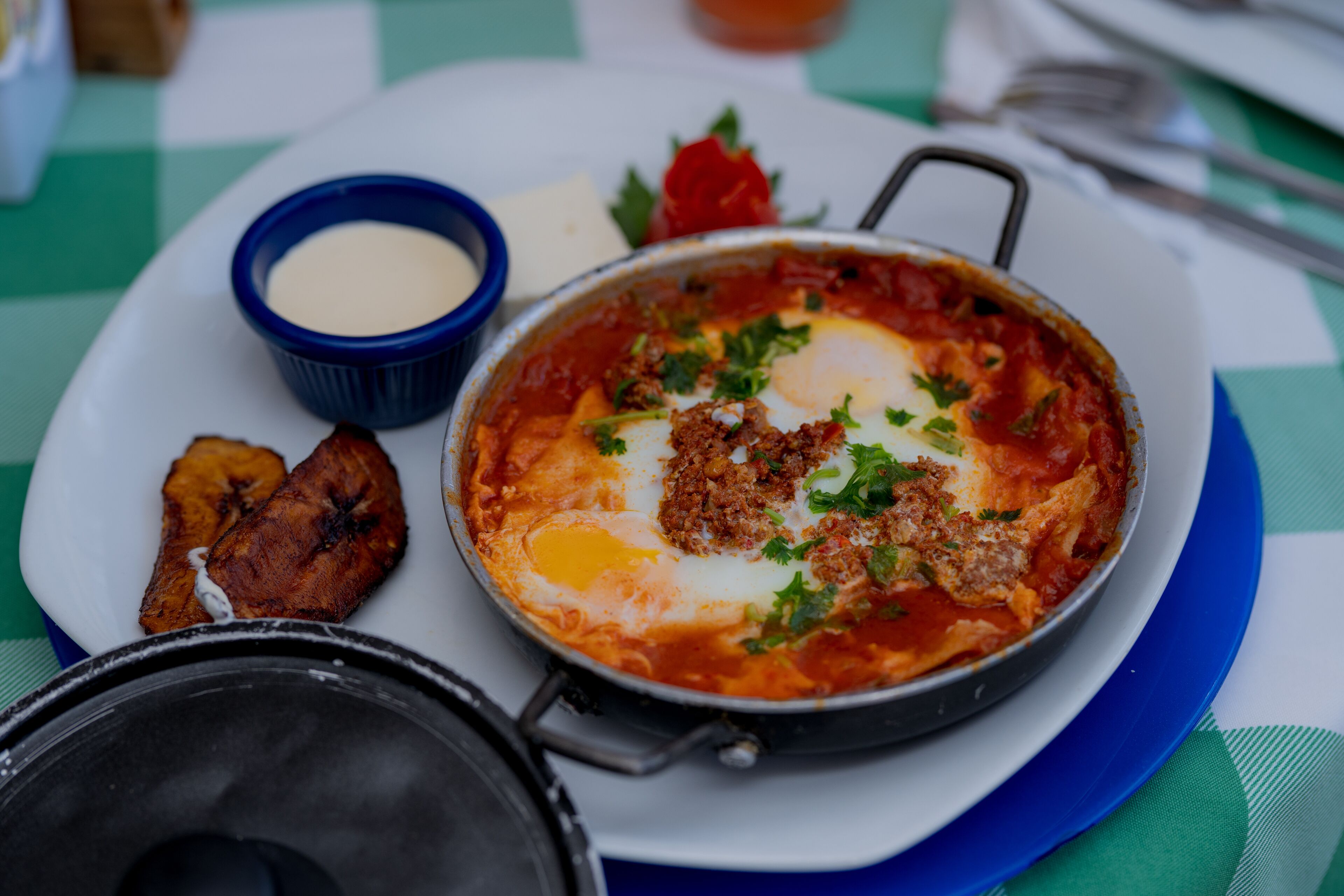 A meal consisting of a pan of eggs with meat and vegetables, a small bowl of sauce, and some fried plantains on a plate.