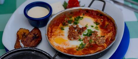 A meal consisting of a pan of eggs with meat and vegetables, a small bowl of sauce, and some fried plantains on a plate.